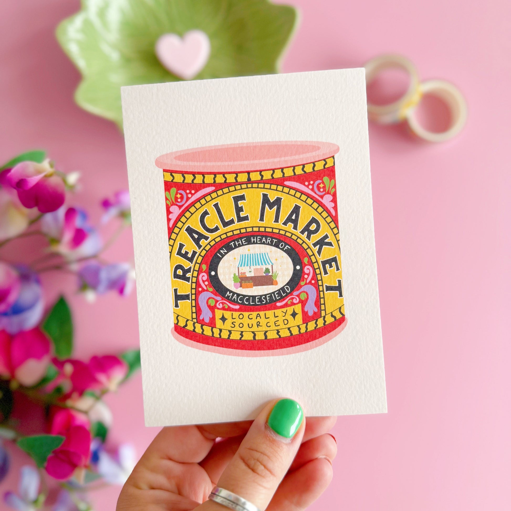 Colourful Treacle Market tin design on a card held by a hand with green nail polish against a pink background.