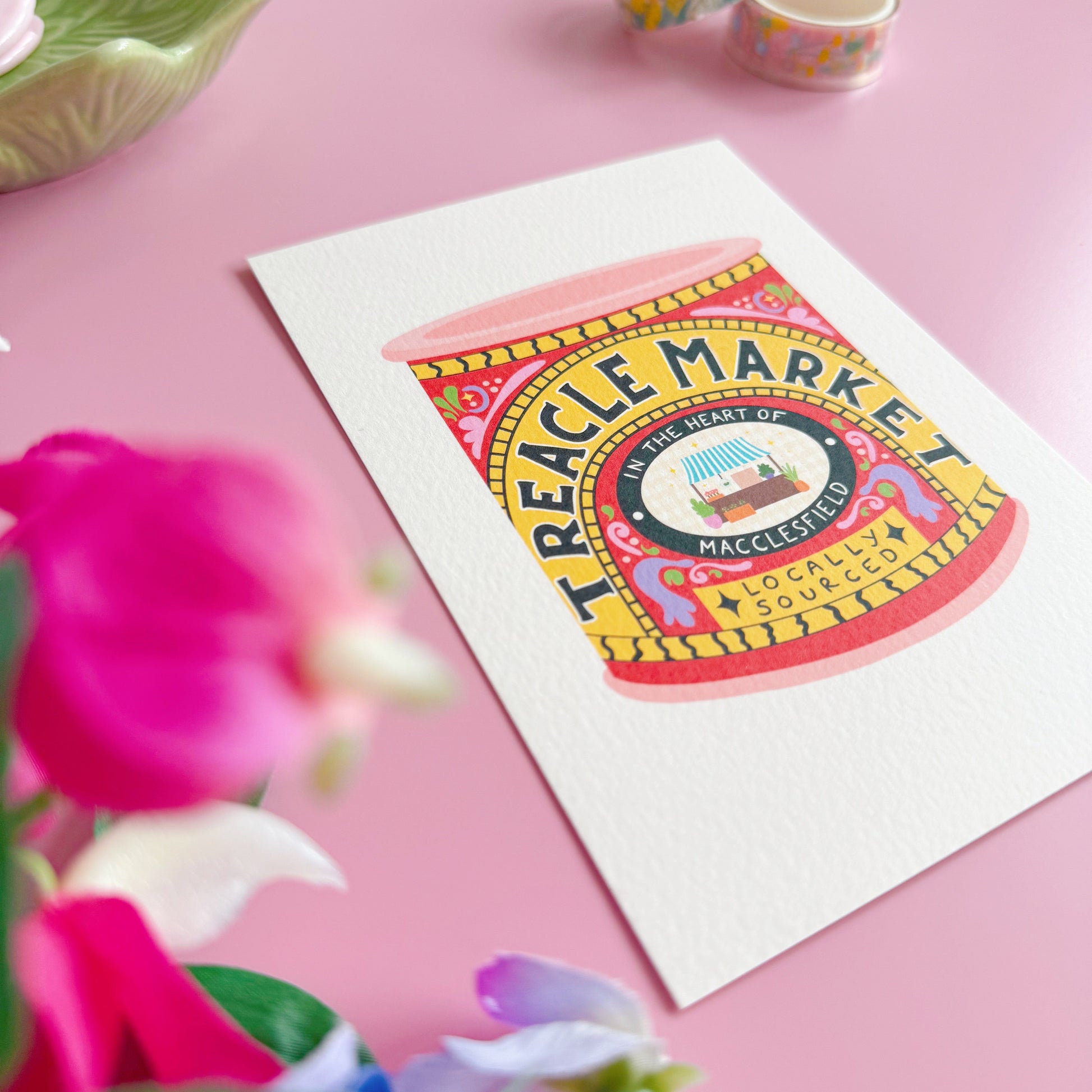 Colorful 'Treacle Market' poster on a pink surface with flowers and small containers.