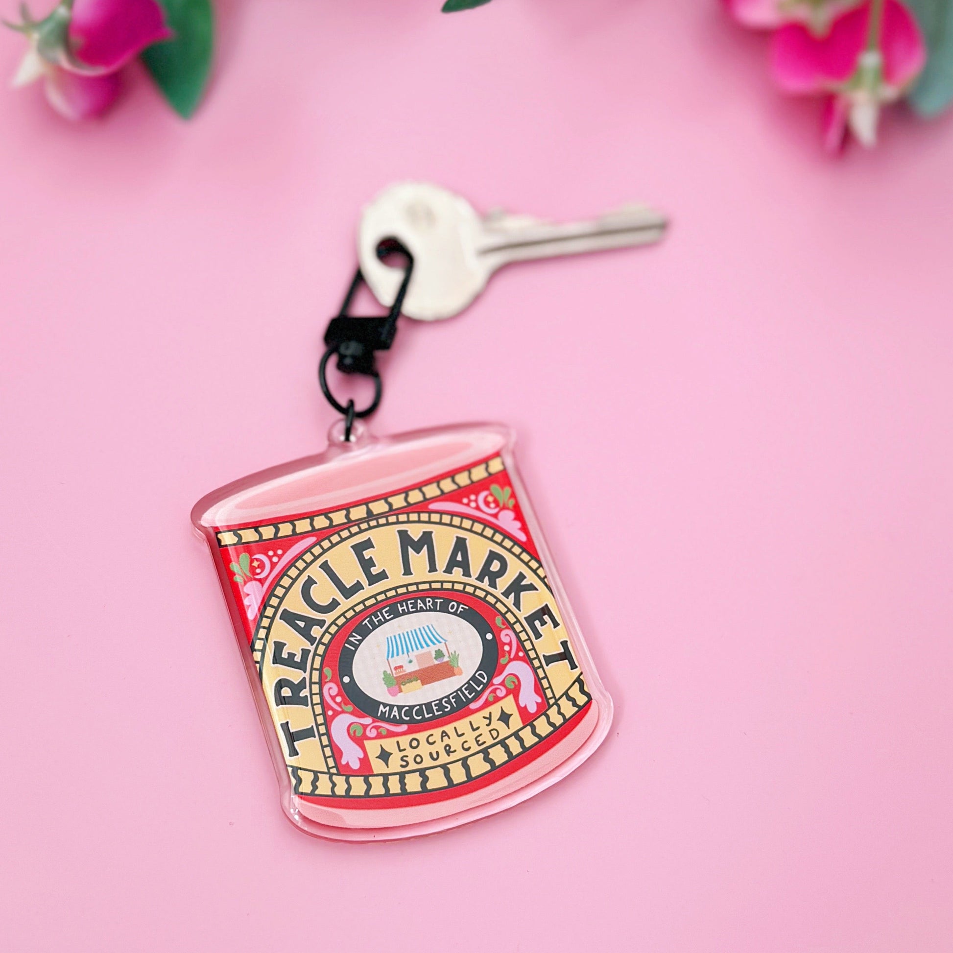 Hand holding a small Treacle Market tin keyring against a pink background with flowers.