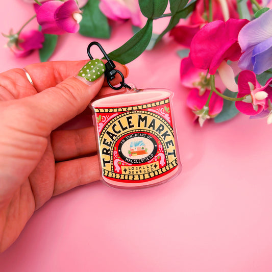 Hand holding a small Treacle Market tin keyring against a pink background with flowers.
