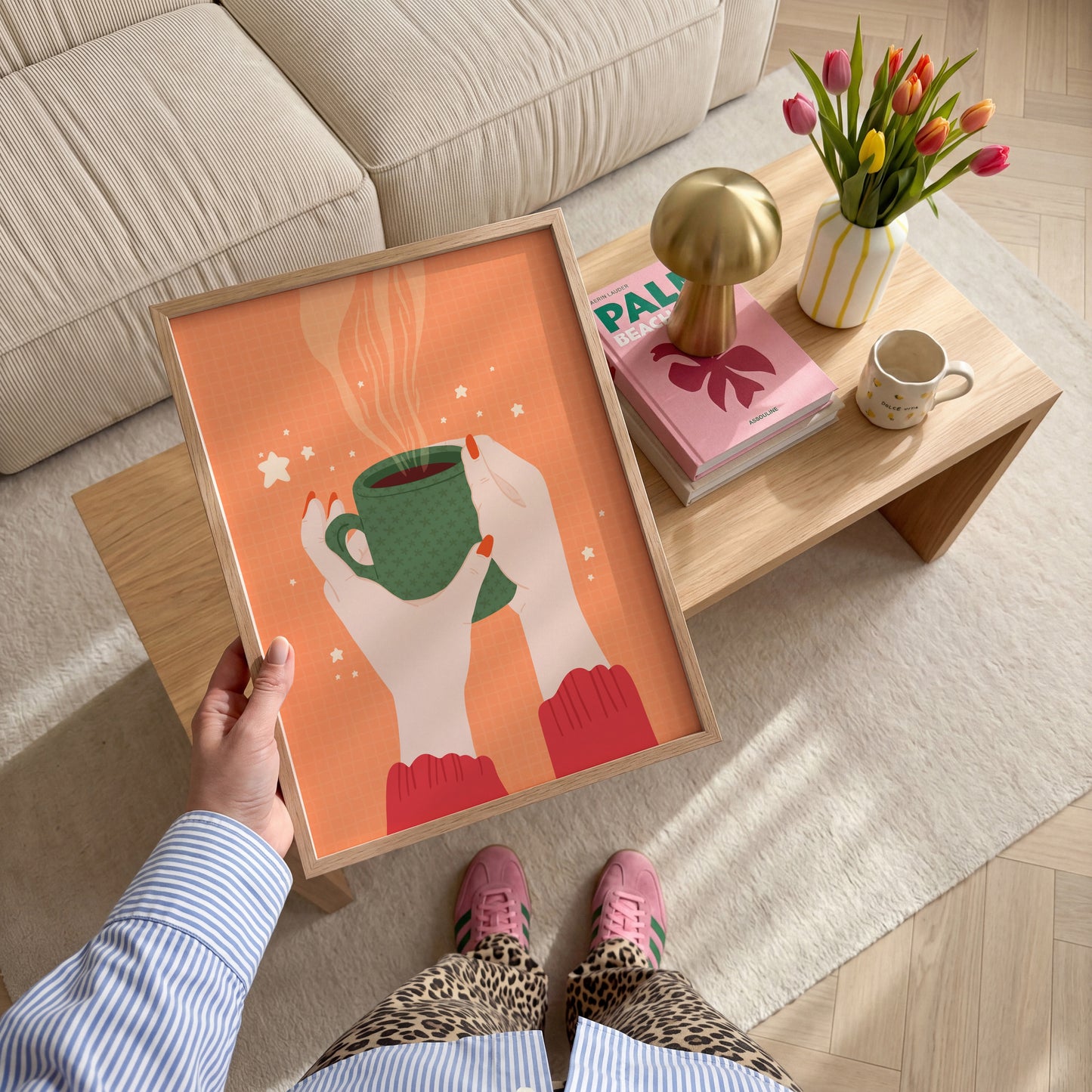 An illustration of a pair of woman's hands holding a green patterned mug, with coffee in. The print background is orange and there are little white stars around the illustration.