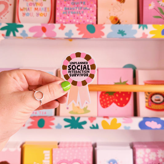 A waterproof vinyl sticker featuring an illustration of an award rosette, with the words 'unplanned social interaction survivor' in the centre.