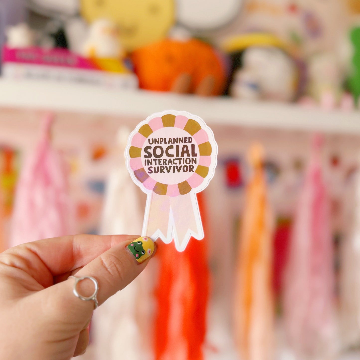 a sticker designed as a rosette, that says 'unplanned social interaction survivor' in pink and green.