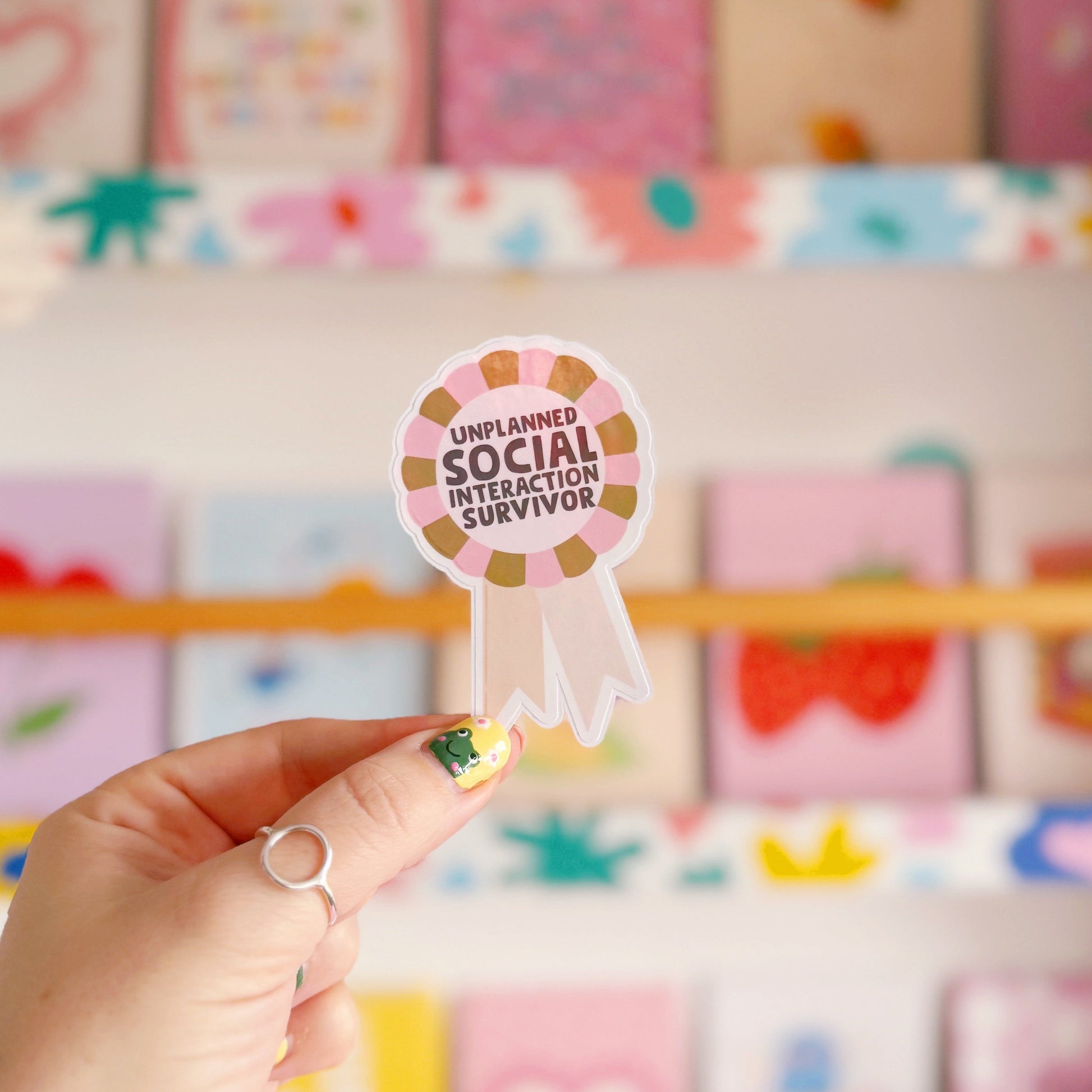 a sticker designed as a rosette, that says 'unplanned social interaction survivor' in pink and green.