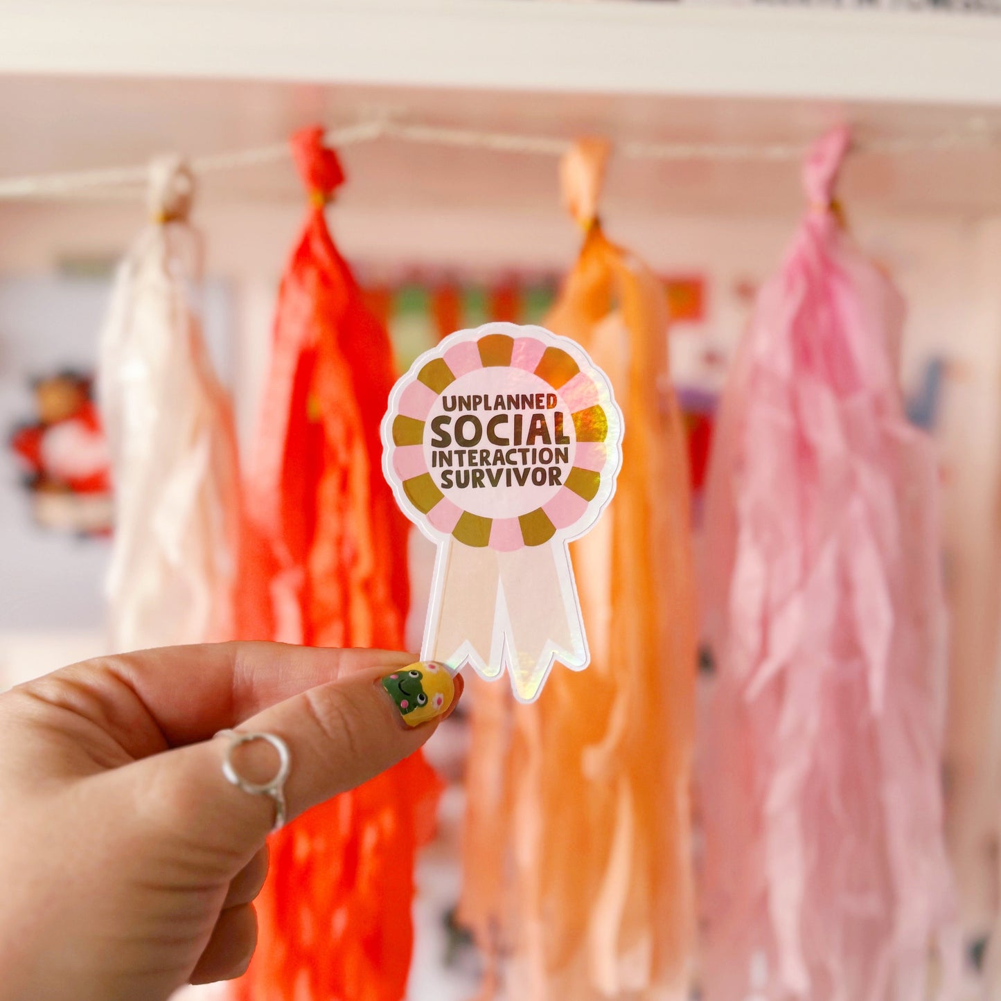 a sticker designed as a rosette, that says 'unplanned social interaction survivor' in pink and green.