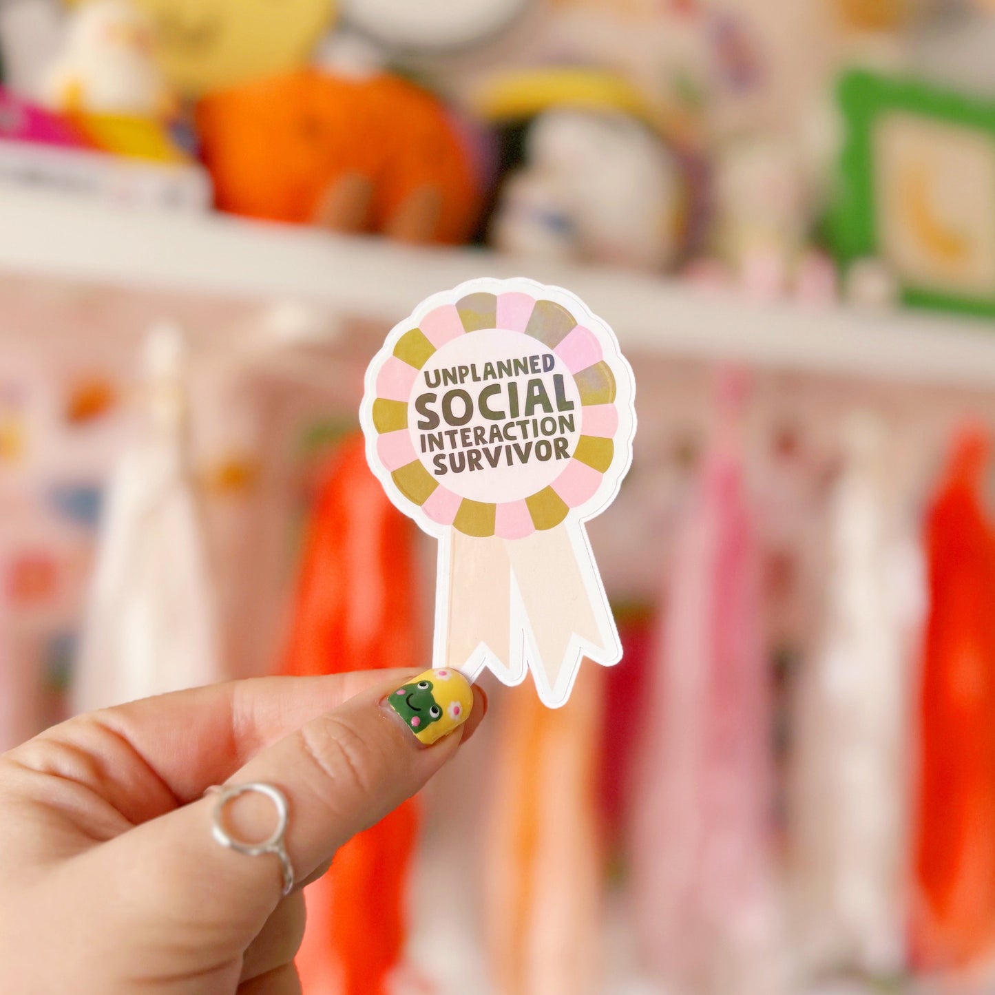 a sticker designed as a rosette, that says 'unplanned social interaction survivor' in pink and green.