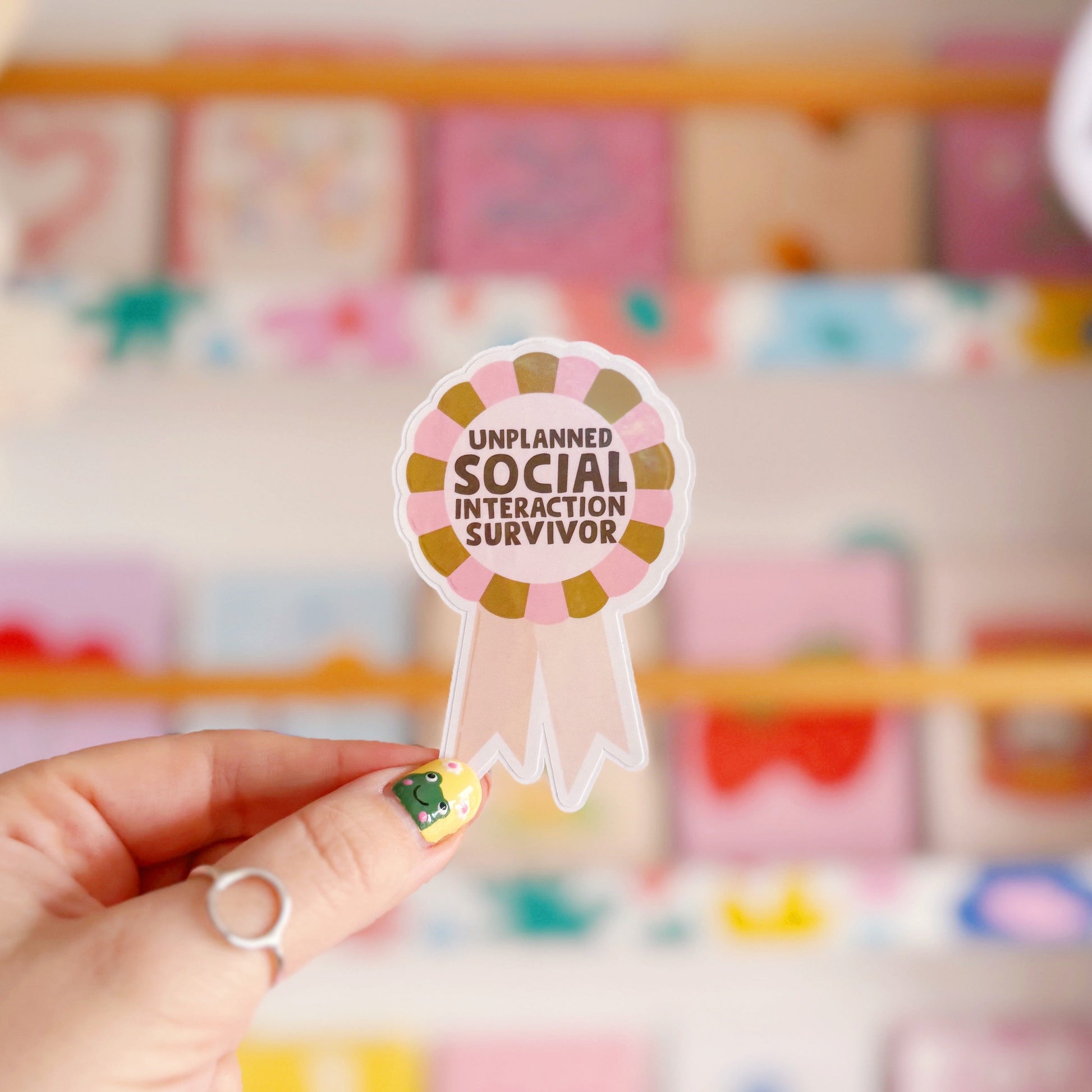 a sticker designed as a rosette, that says 'unplanned social interaction survivor' in pink and green.