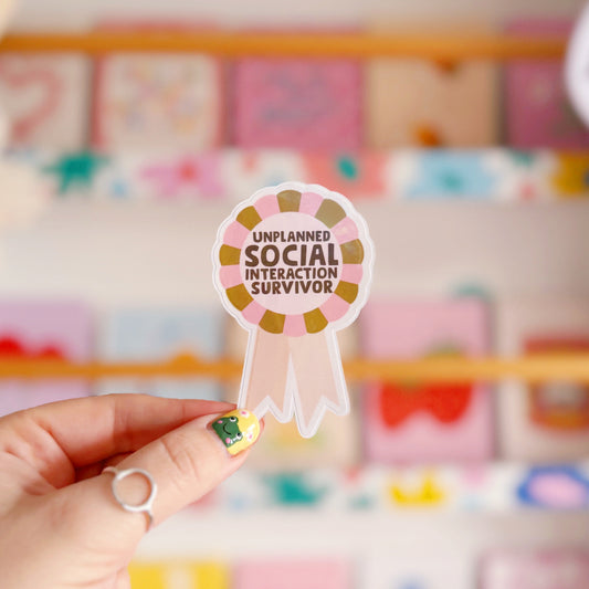 a sticker designed as a rosette, that says 'unplanned social interaction survivor' in pink and green.