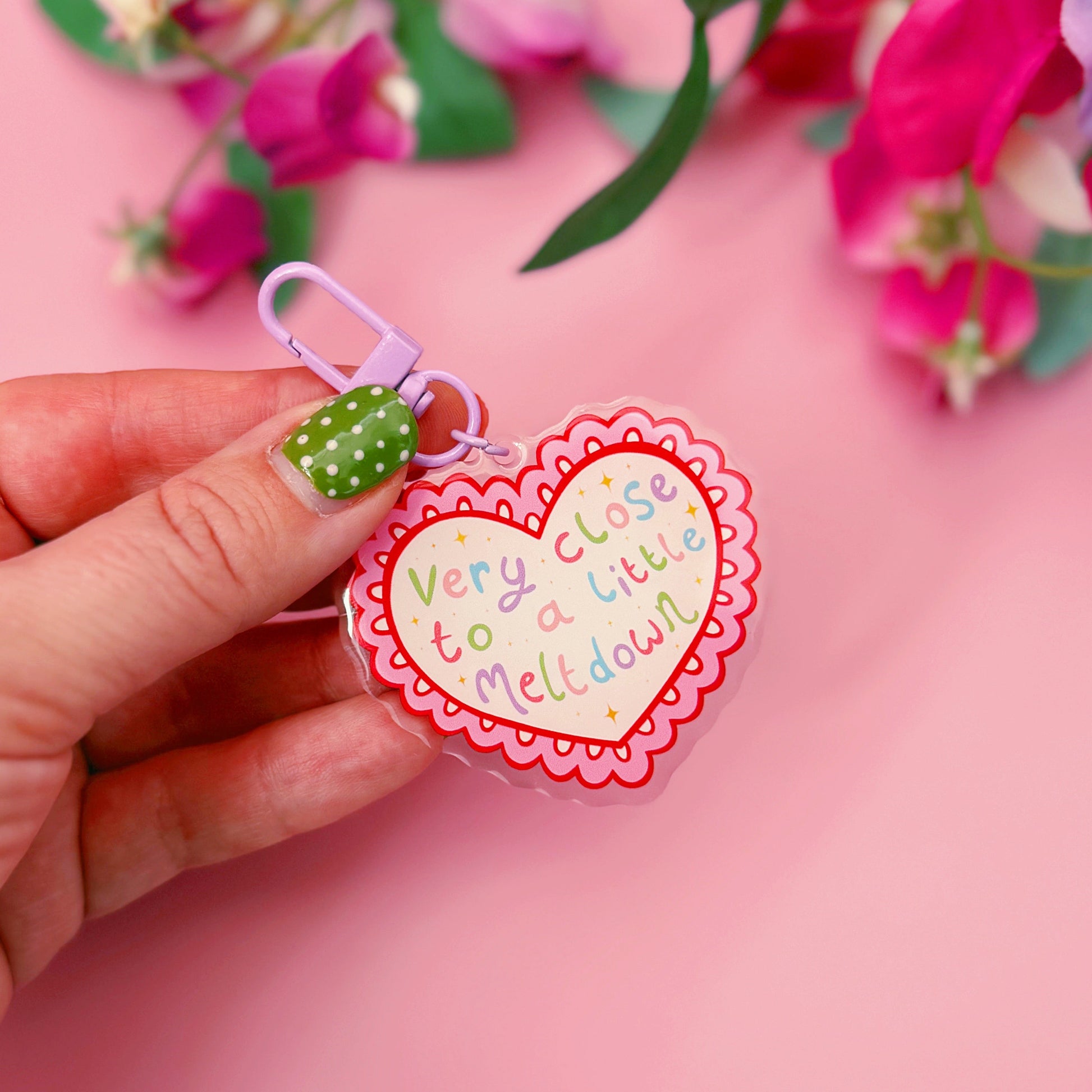 Heart-shaped keychain with text held by a hand against a pink background with flowers.