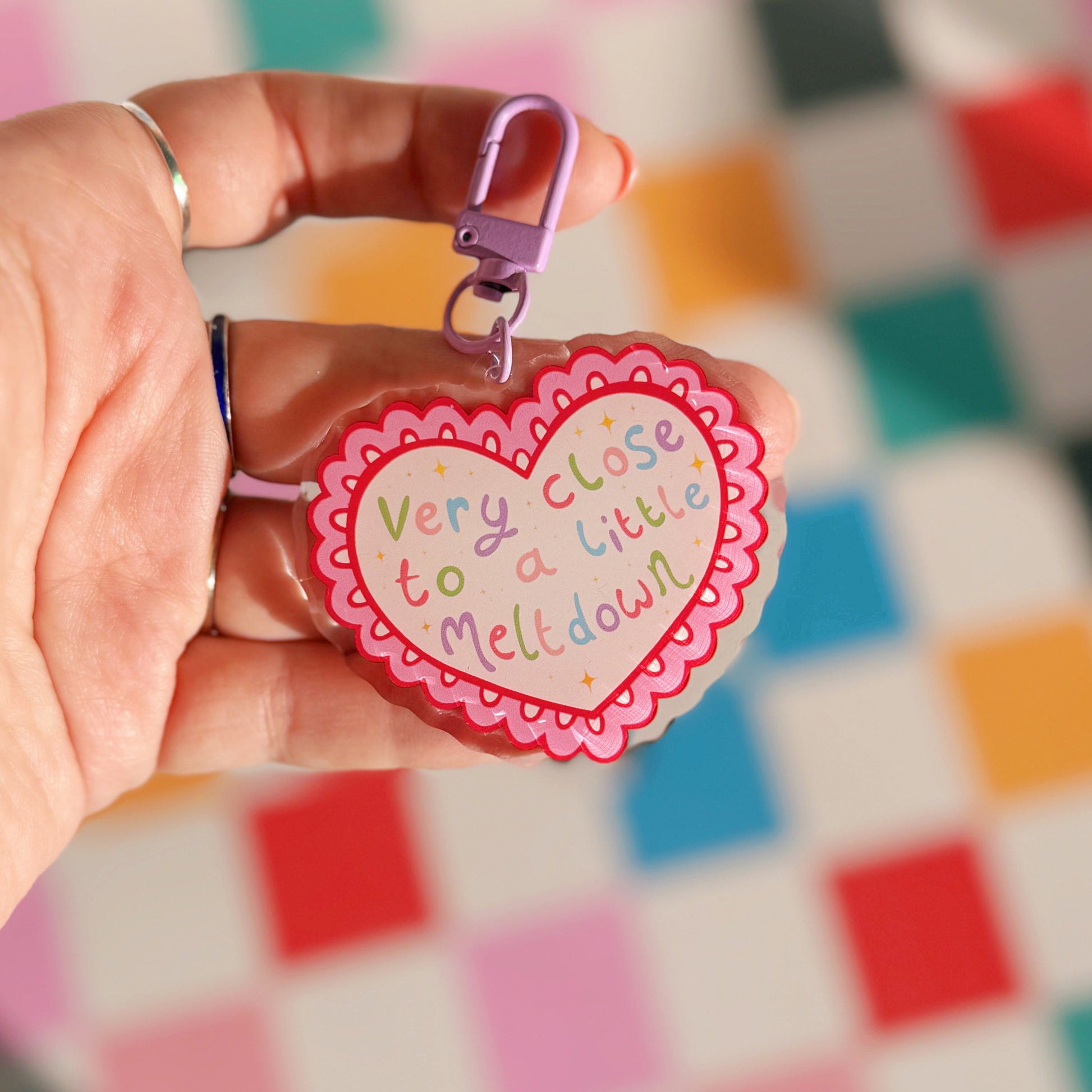 Heart-shaped keychain with text held by a hand against a colourful checkered background