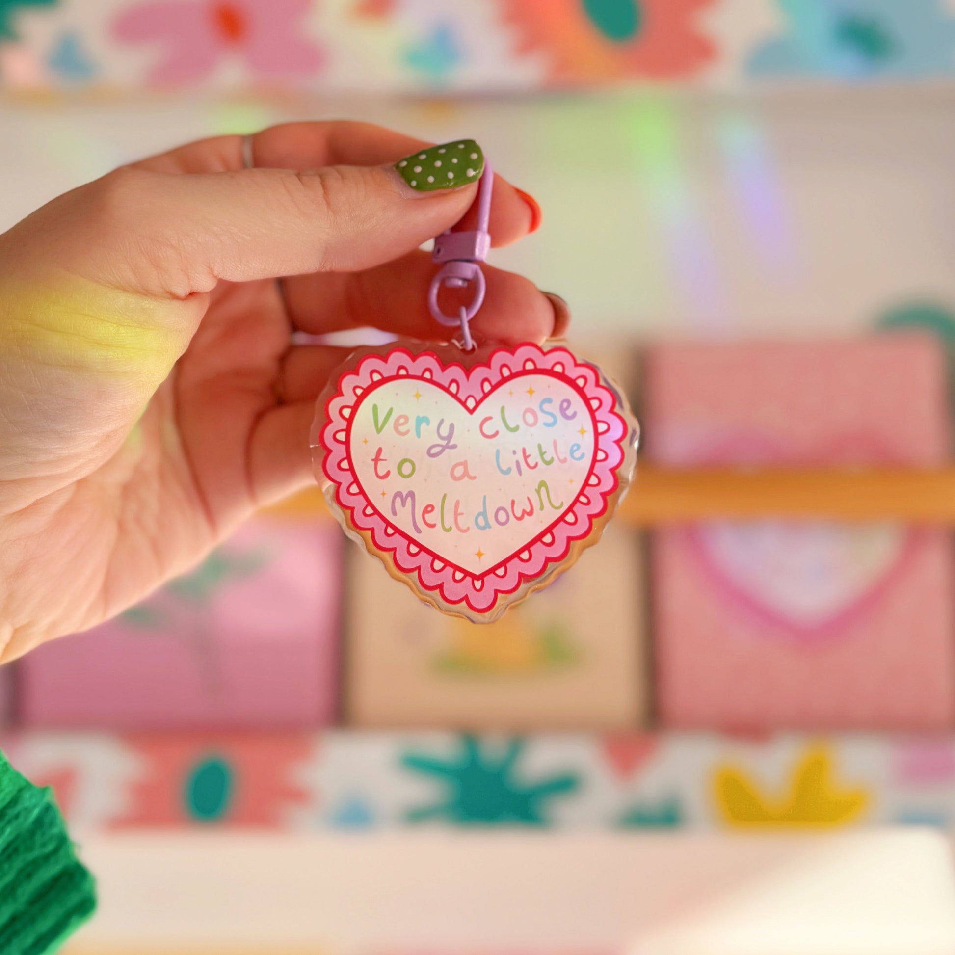Heart-shaped keychain with 'Very close to a little Meltdown' text held by a hand against a colourful background.