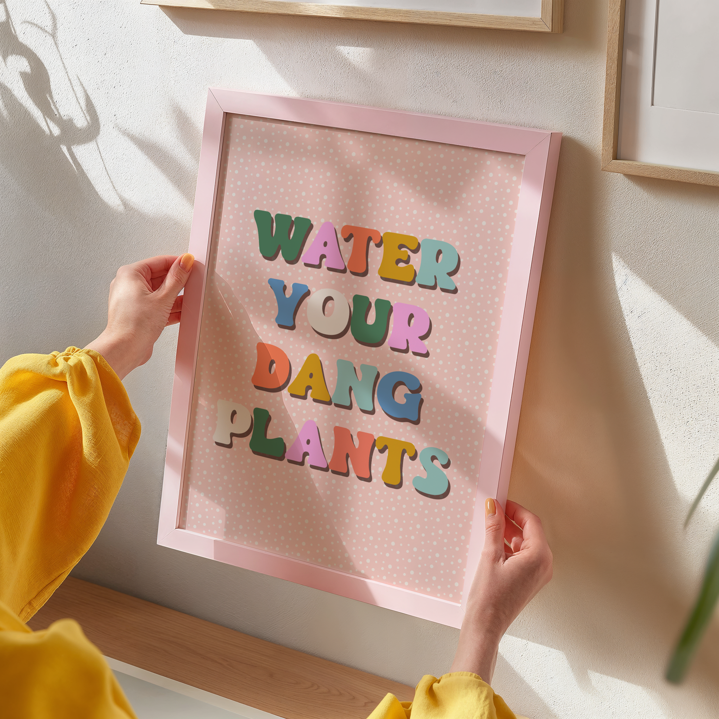 A quote print, saying water your dang plants, in multicoloured text on a peachy pink background.