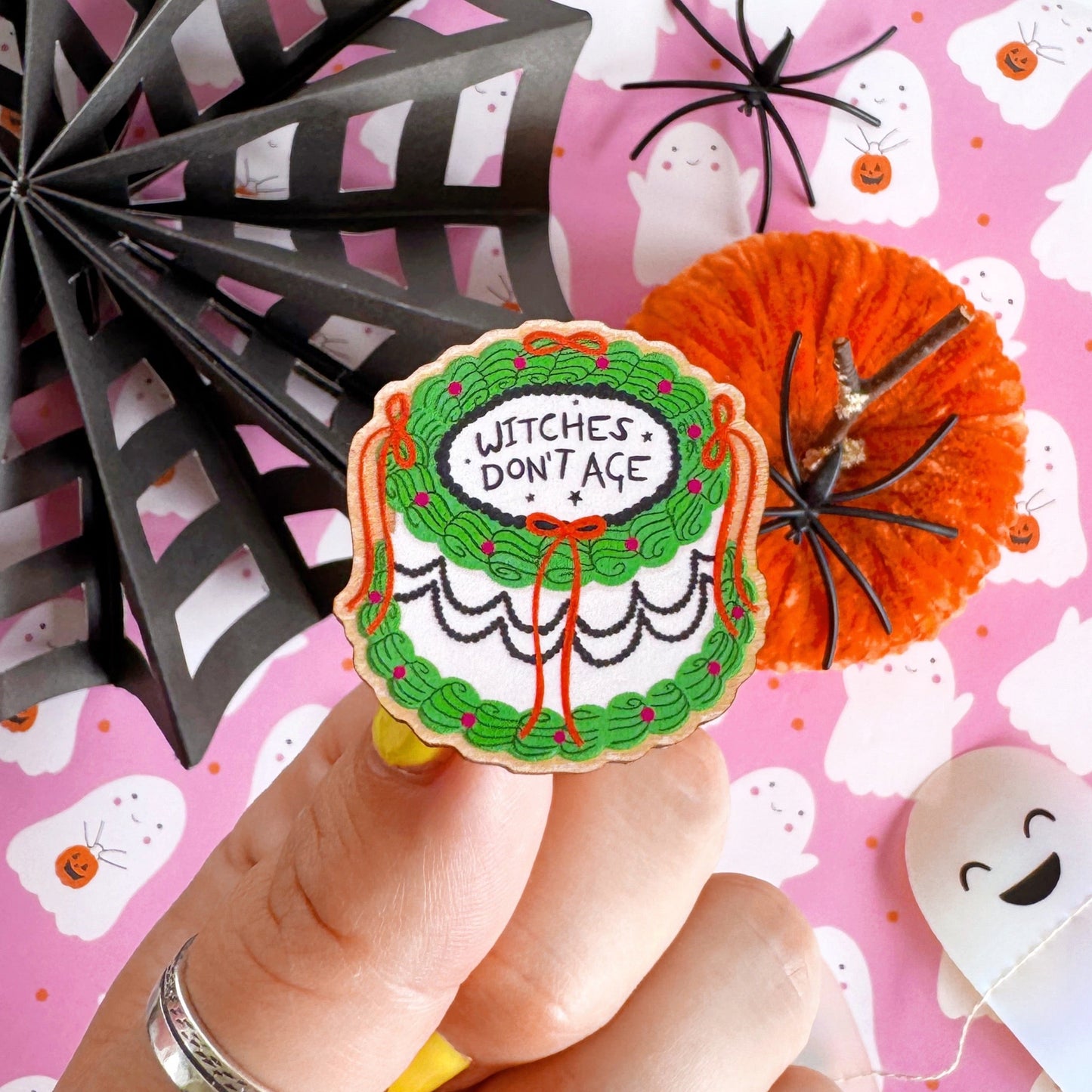 A halloween themed cake pin badge, with green white and black icing and orange bows. The text on the top of the cake reads 'Witches don't age'.