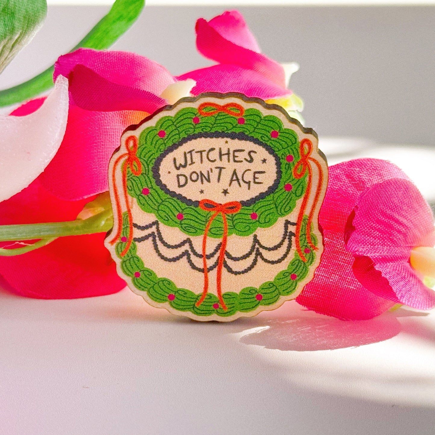A cake shaped wooden pin badge, in green black and orange, with lack icing that says witches don't age.