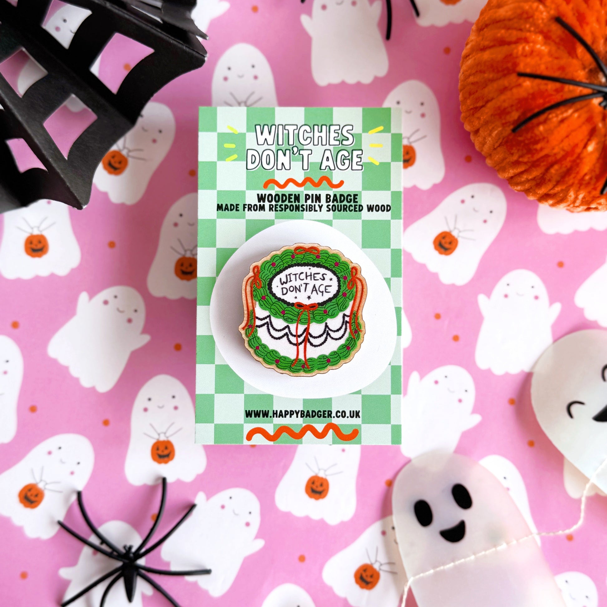A halloween themed cake pin badge, with green white and black icing and orange bows. The text on the top of the cake reads 'Witches don't age'.