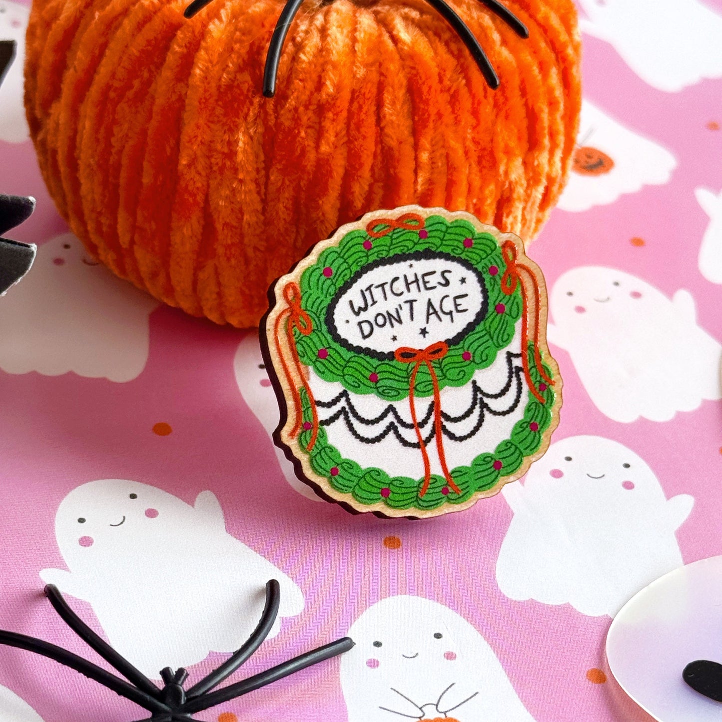 A halloween themed cake pin badge, with green white and black icing and orange bows. The text on the top of the cake reads 'Witches don't age'.
