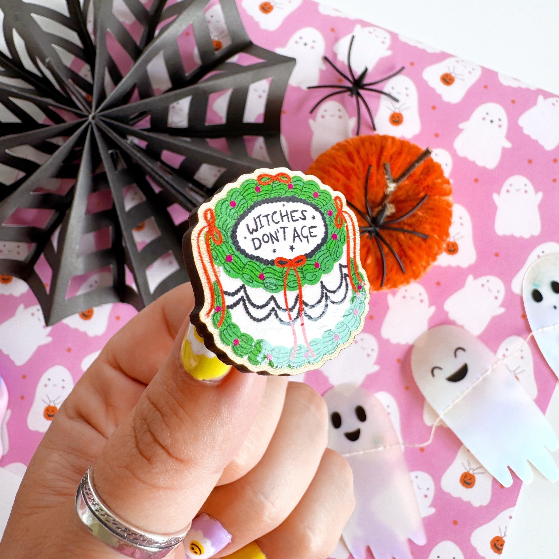 A halloween themed cake pin badge, with green white and black icing and orange bows. The text on the top of the cake reads 'Witches don't age'.