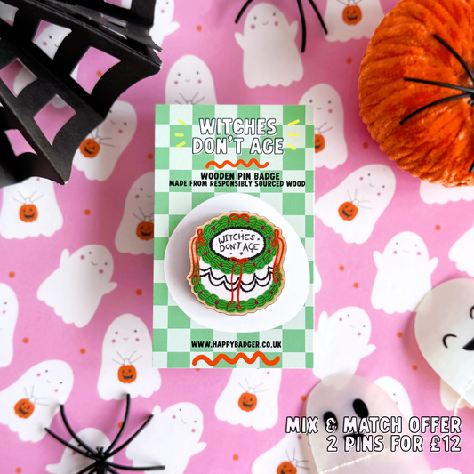 A halloween themed cake pin badge, with green white and black icing and orange bows. The text on the top of the cake reads 'Witches don't age'.