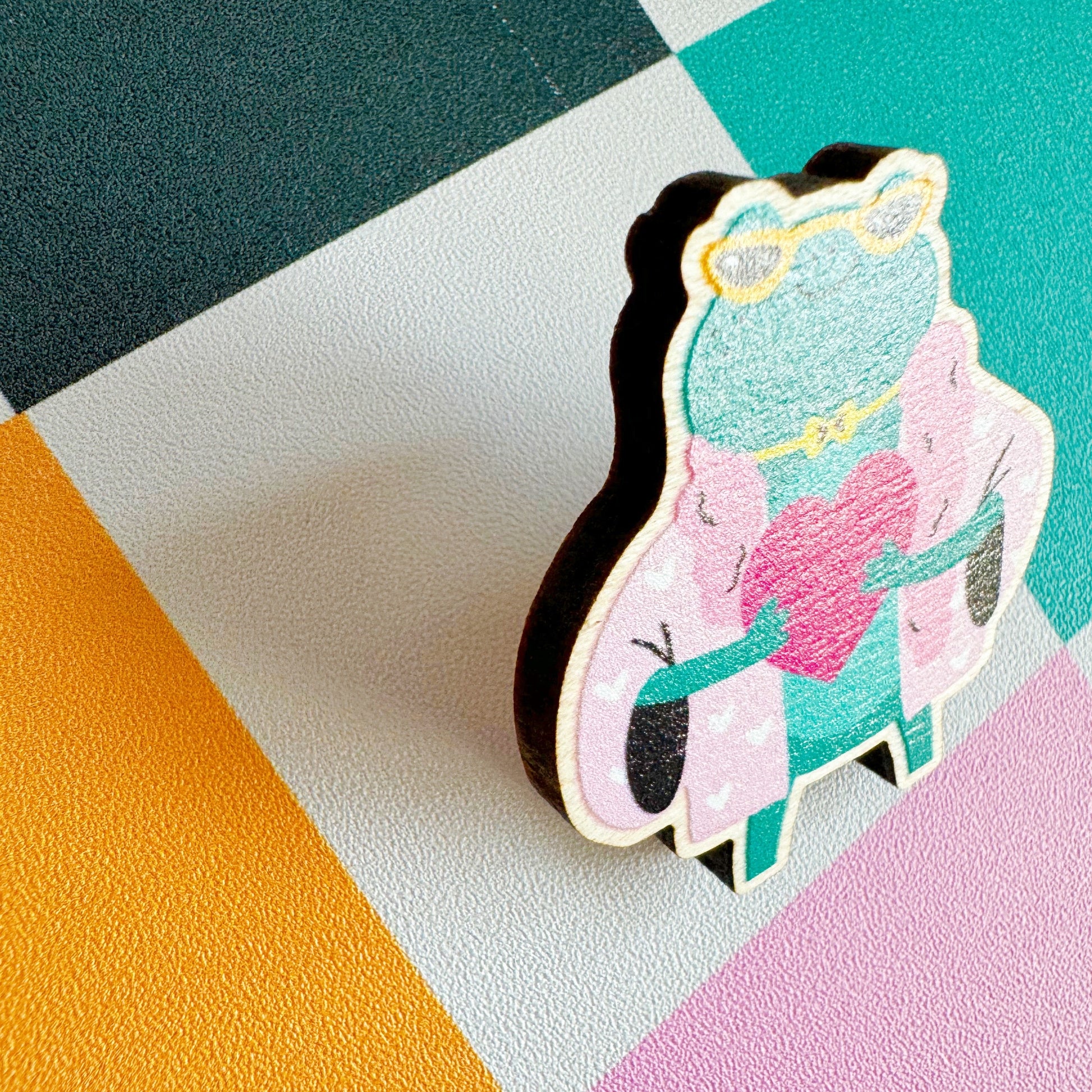 A cute illustration of a frog in sunglasses and a pink coat, holding a love heart, on an eco friendly wooden pin badge with two back fastenings.