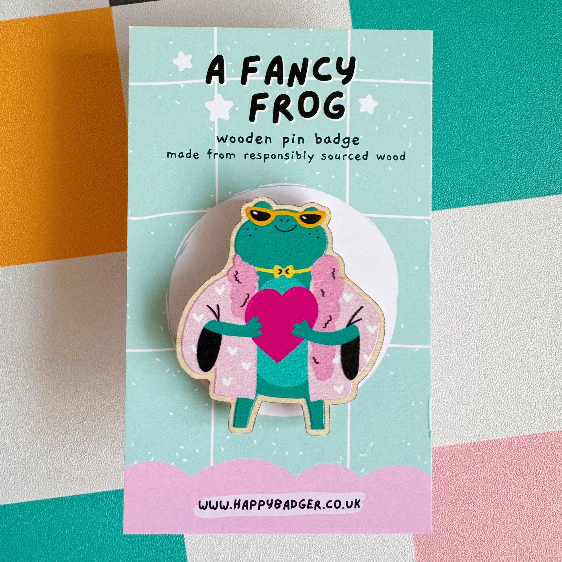 A cute illustration of a frog in sunglasses and a pink coat, holding a love heart, on an eco friendly wooden pin badge with two back fastenings.