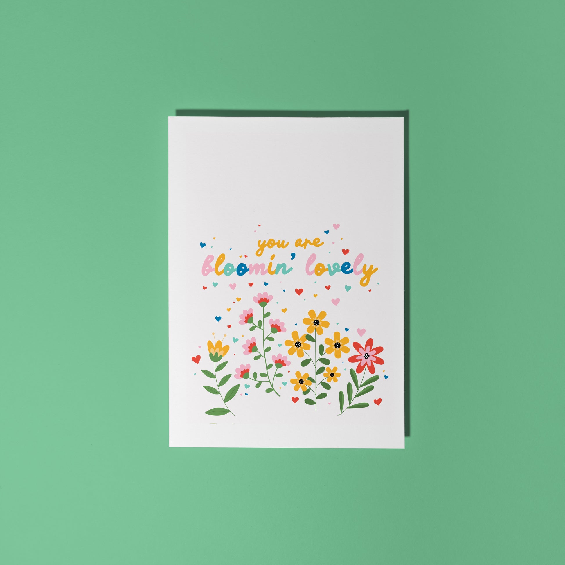 A white card, with a floral illustration scene at the bottom and the words 'you are bloomin' lovely' in colourful handwriting above the flowers.