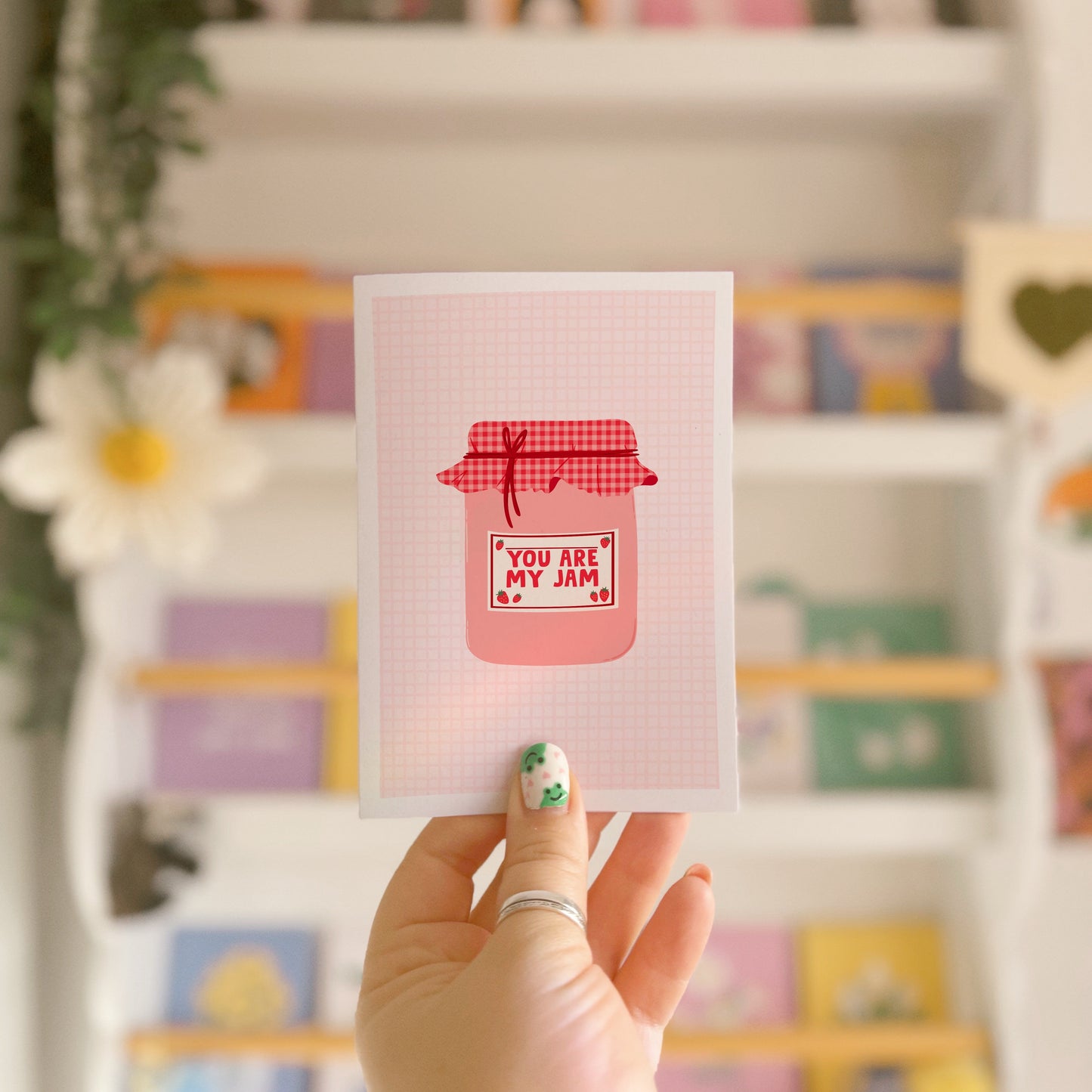 A greeting card with an illustration of a jam jar, with 'you are my jam' written on the label. The jam jar has a red and pink gingham lid and the label is decorated with strawberry doodles.