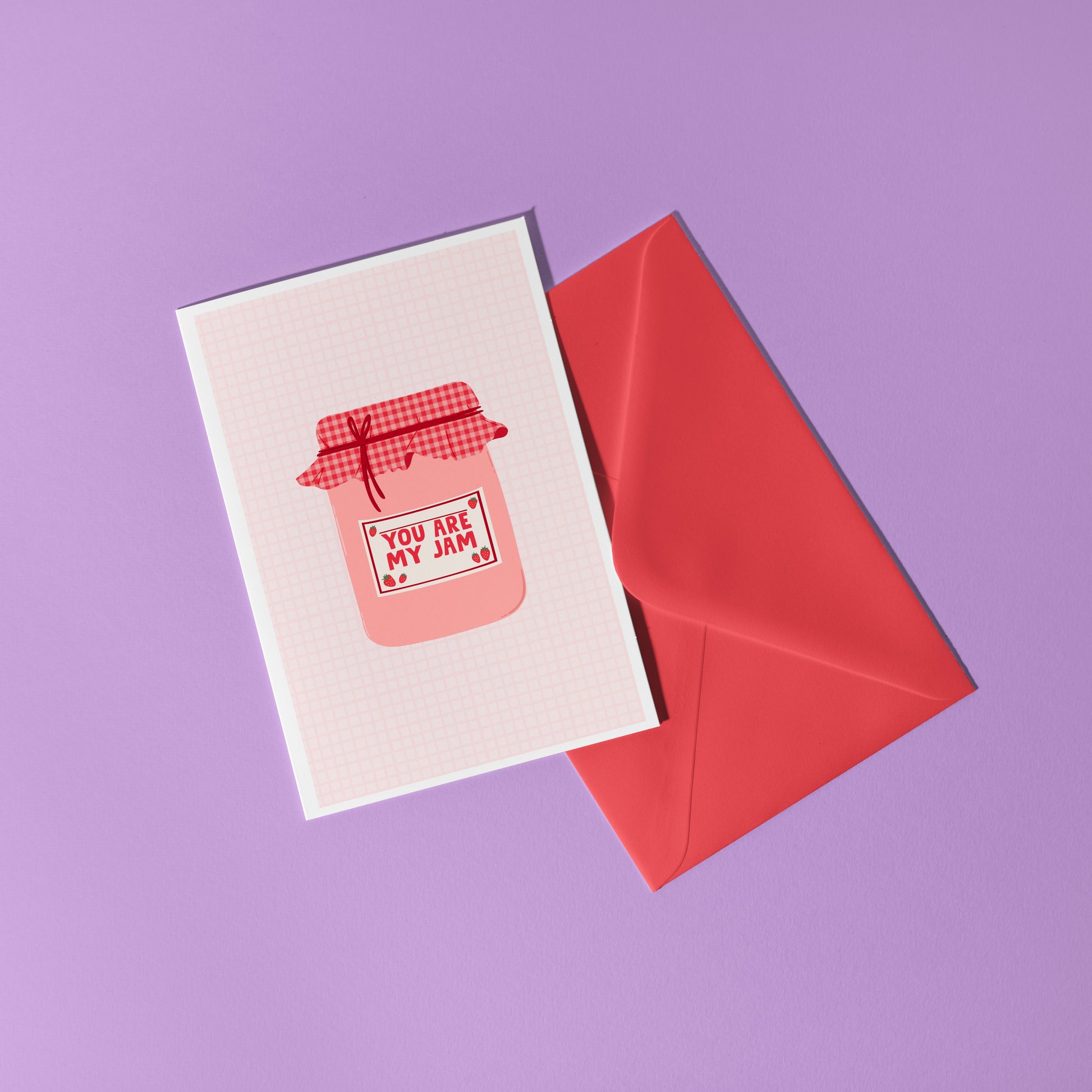 A greeting card with an illustration of a jam jar, with 'you are my jam' written on the label. The jam jar has a red and pink gingham lid and the label is decorated with strawberry doodles.