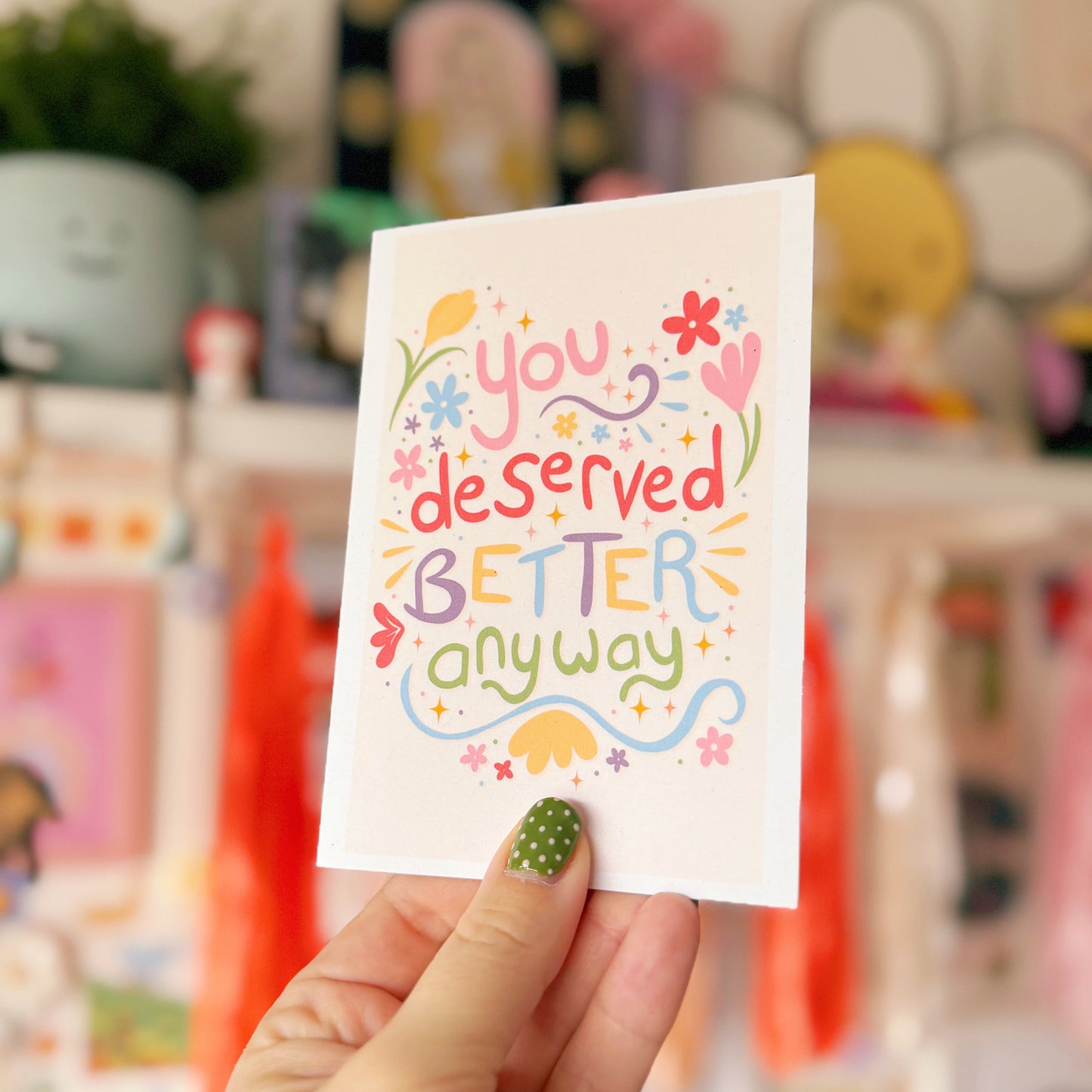 Hand holding a card with 'You deserved better anyway' text in colorful letters against a blurred background.