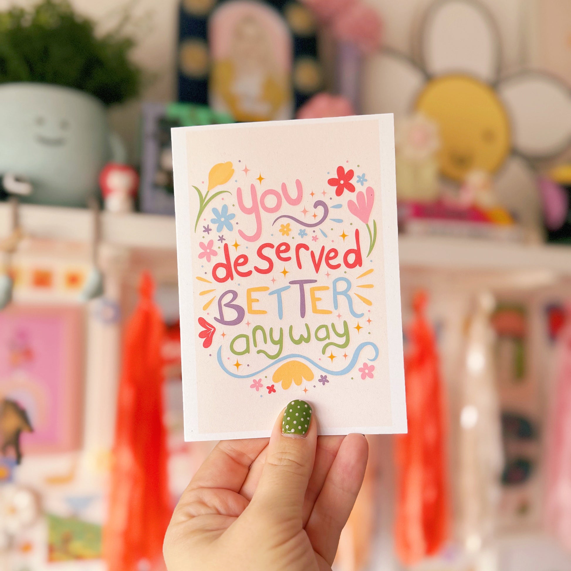 Hand holding a card with 'you deserved better anyway' text in a colorful font against a blurred store background.