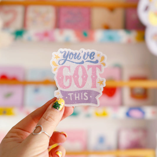 A holographic sticker with script writing saying 'you've got this' in blue, pink and purple, with little yellow illustrated detailing around the text.