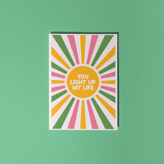 A pink, green and yellow star burst design with the words 'you light up my life' in the centre.