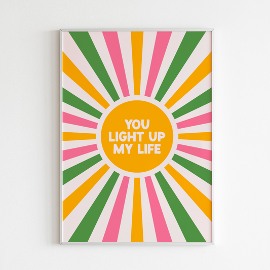 A bright vibrant print, showing a sunburst pattern in pink green and yellow, with a yellow circle in the middle saying 'You Light Up My Life'.