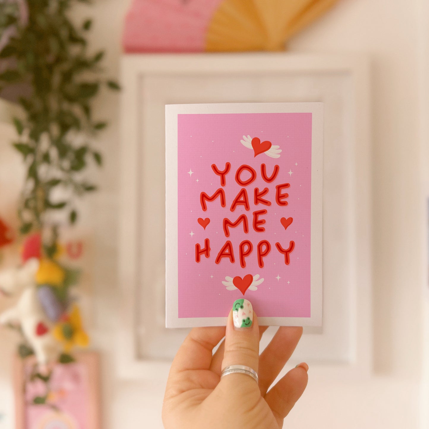 A pink greeting card with little heart illustrations around a red hand lettered quote that says 'you make me happy'.