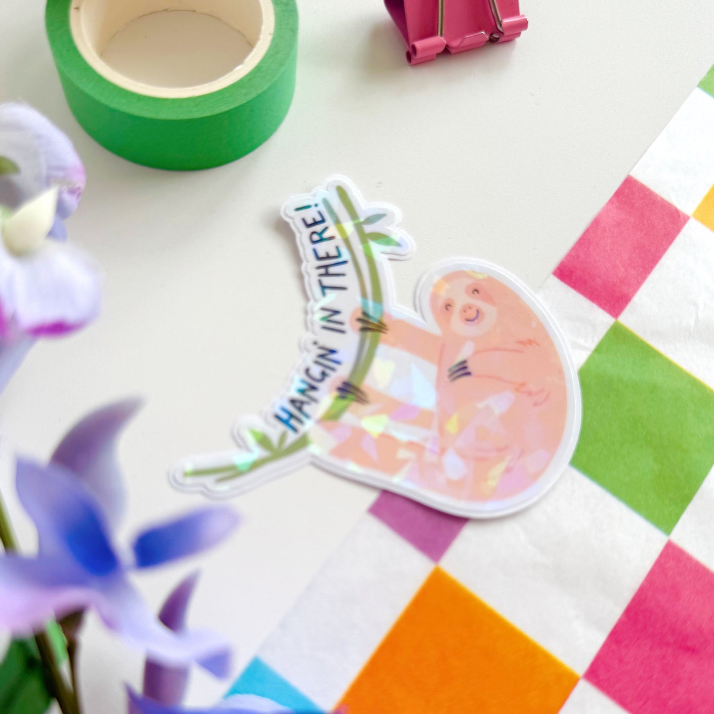 A holographic sticker with a sloth and 'Hangin' in There!' text.