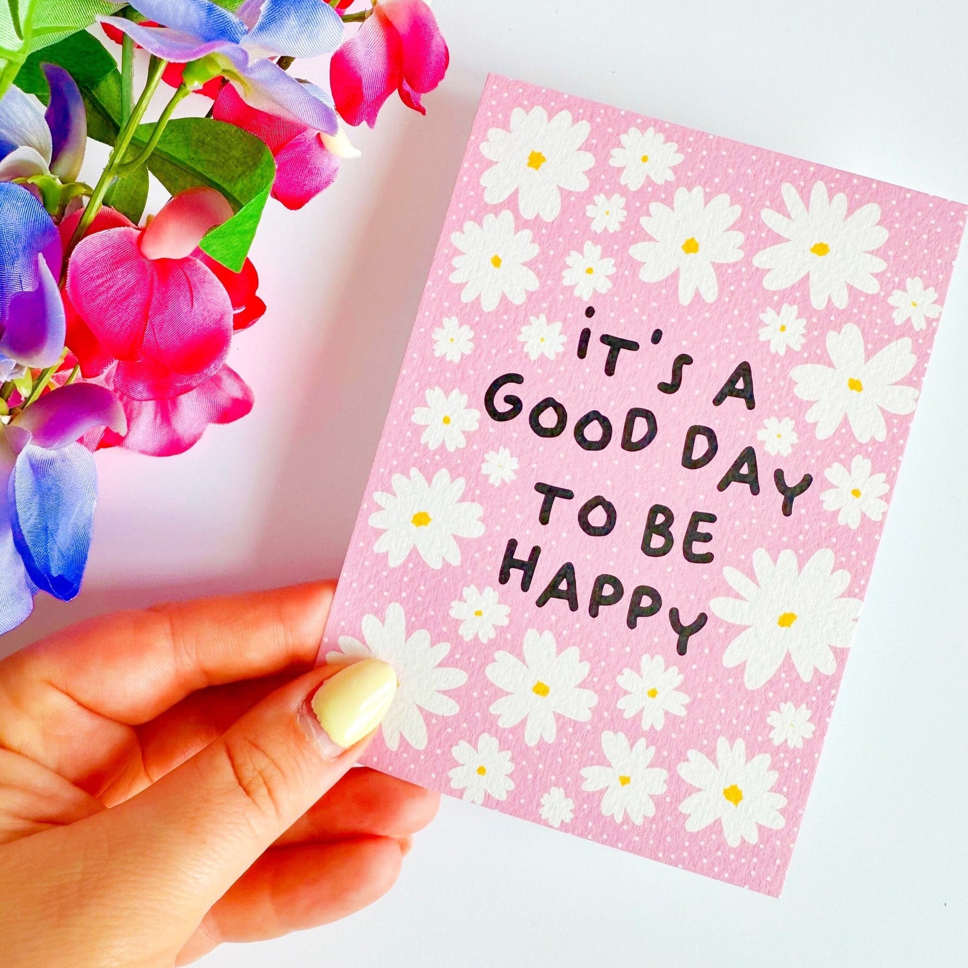 A textured postcard, with the words it's a good day to be happy, on the front, on a pink background with white daises.