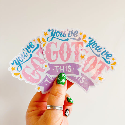 A hand holding three glittery stickers, that say 'You've Got This' in pink blue and purple colours.