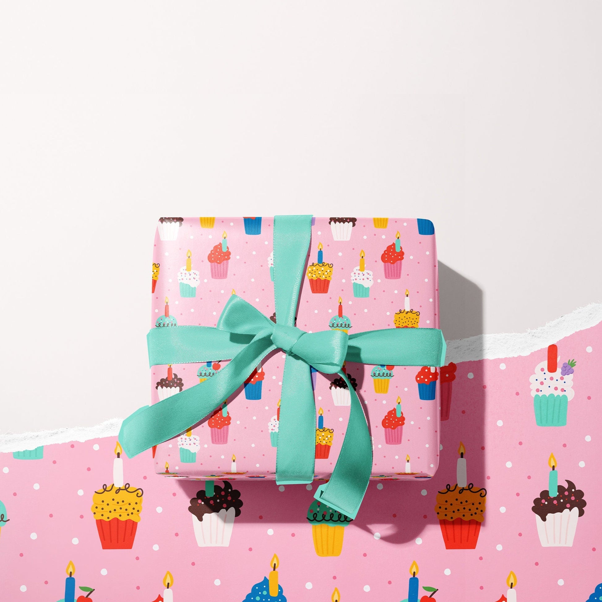 A present wrapped in pink gift wrap, which has colourful cupcakes and candles all over. The present is wrapped with a teal bow.