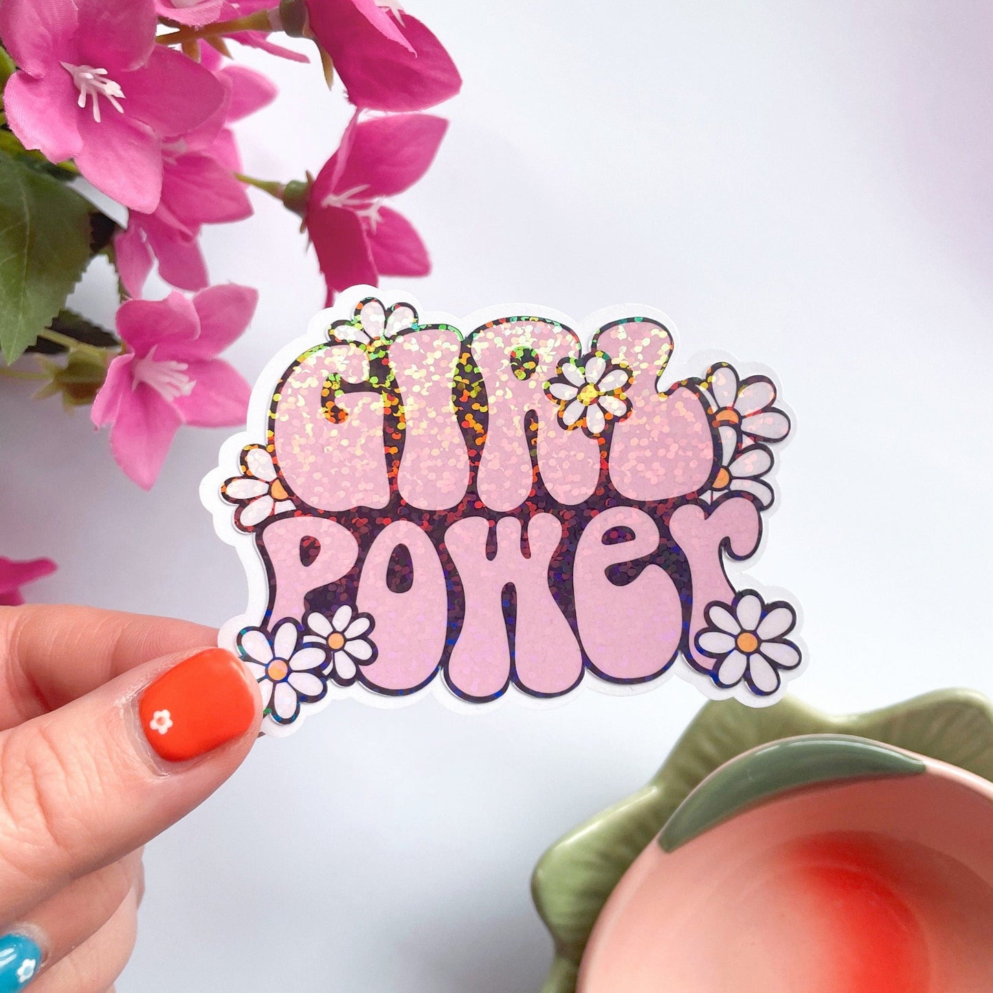 A holographic sticker, of the words girl power in pink bubble writing surrounded by little daisies. The sticker has an easy peel lip.