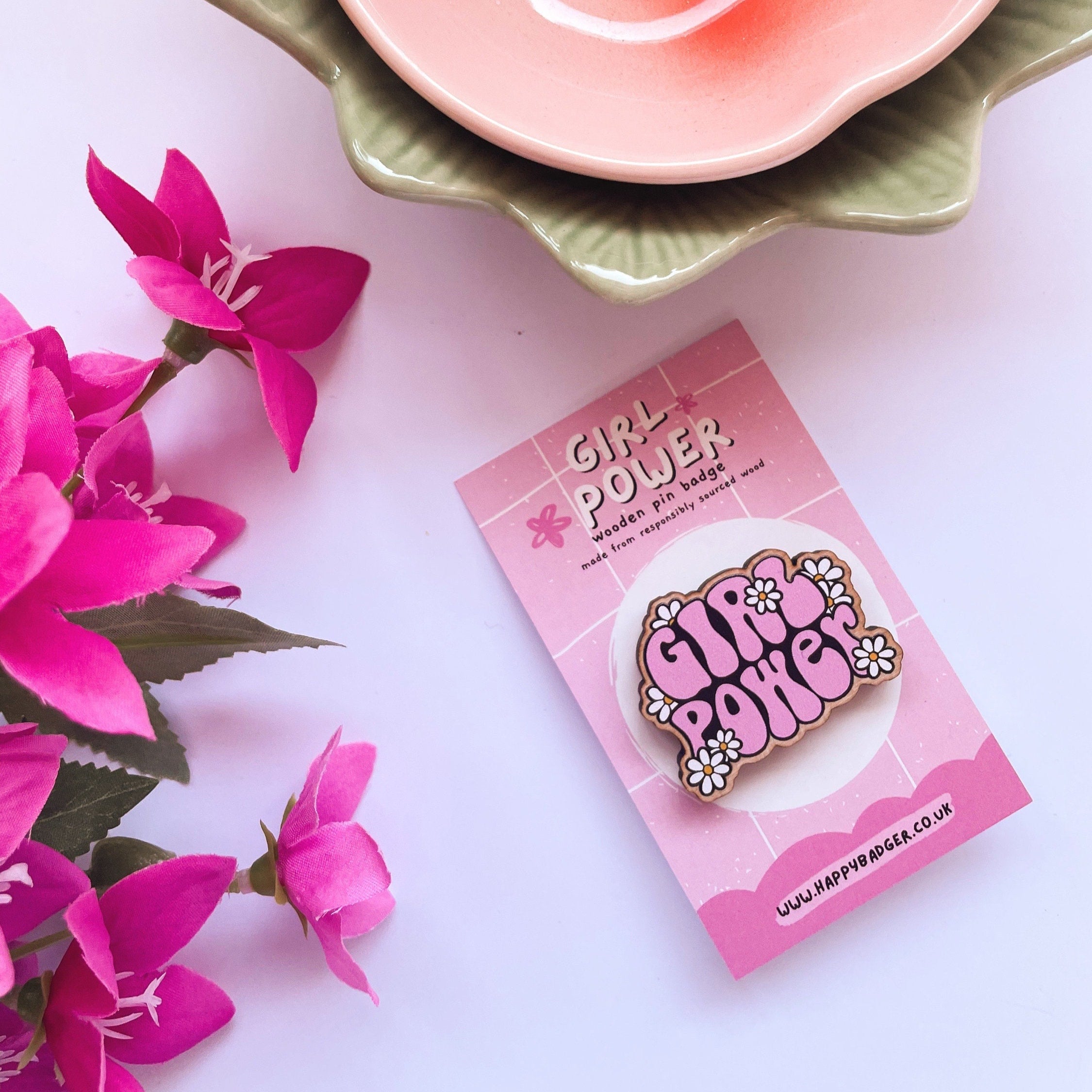 A wooden eco friendly pin badge saying girl power in pink writing, with white daisies around it.