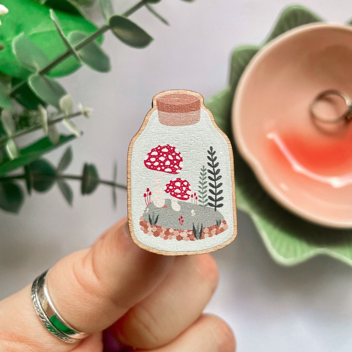 An eco friendly wooden pin badge, of a terrarium with greenery and toadstools inside. The pin badge has two back fastenings.