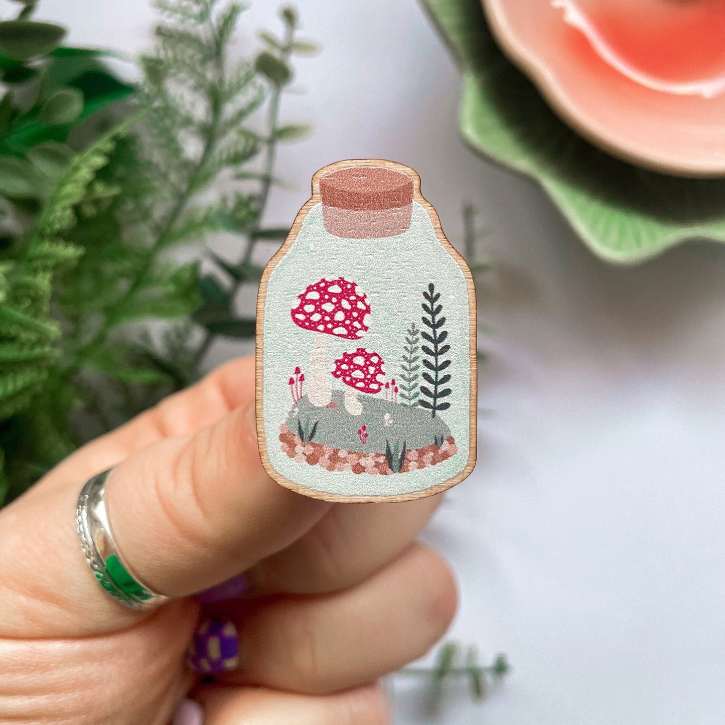 An eco friendly wooden pin badge, of a terrarium with greenery and toadstools inside. The pin badge has two back fastenings.