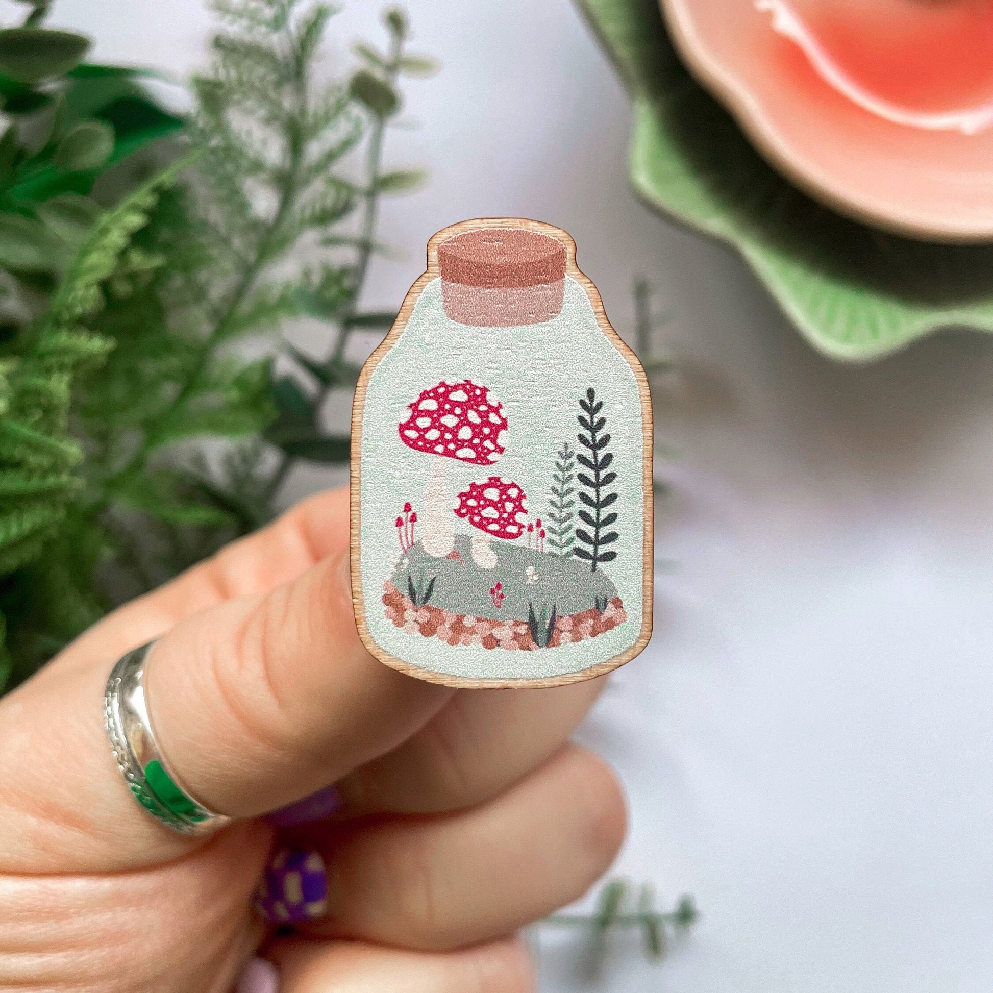 An eco friendly wooden pin badge, of a terrarium with greenery and toadstools inside. The pin badge has two back fastenings.