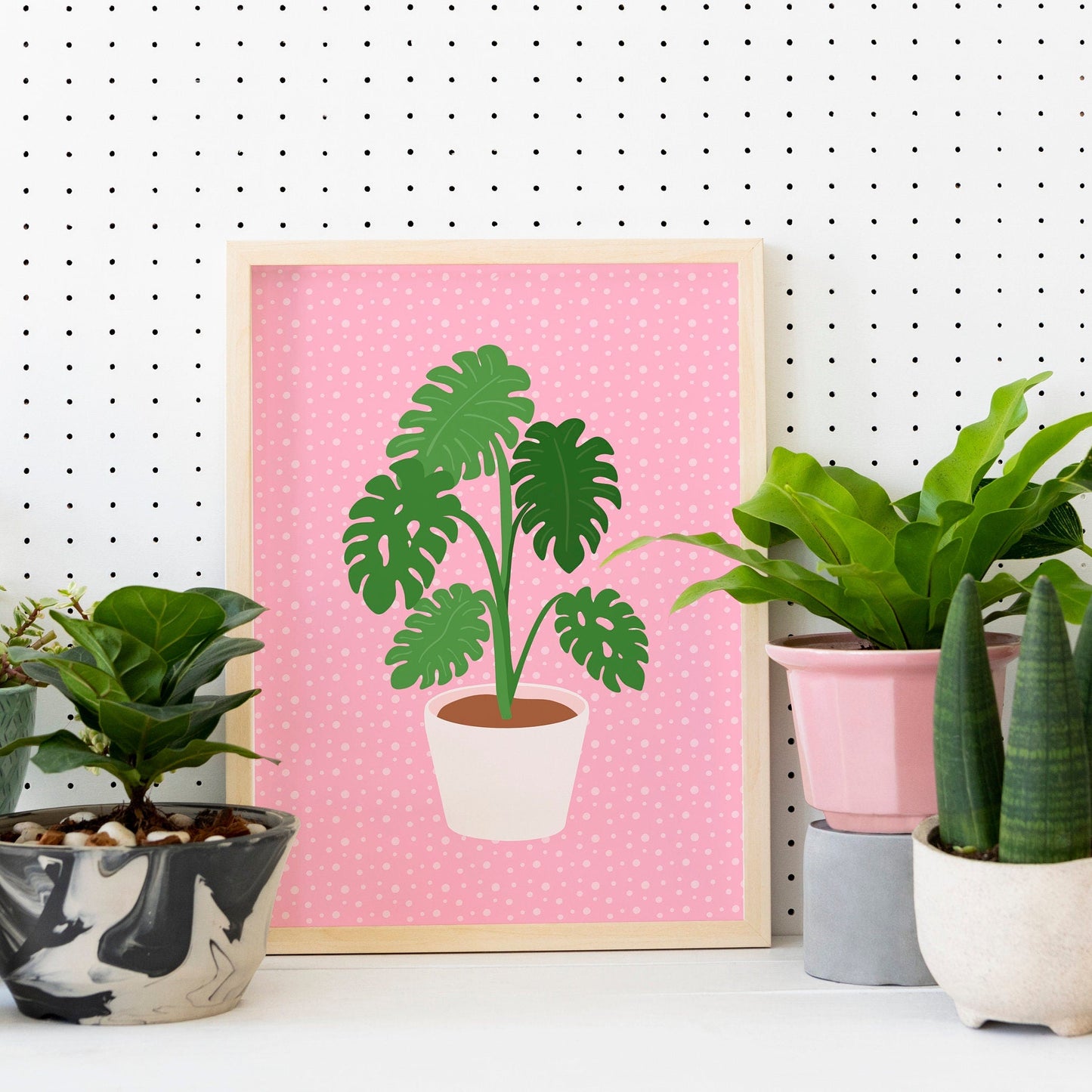A framed print of a Monstera plant in a pot, with green leaves on a pink background.