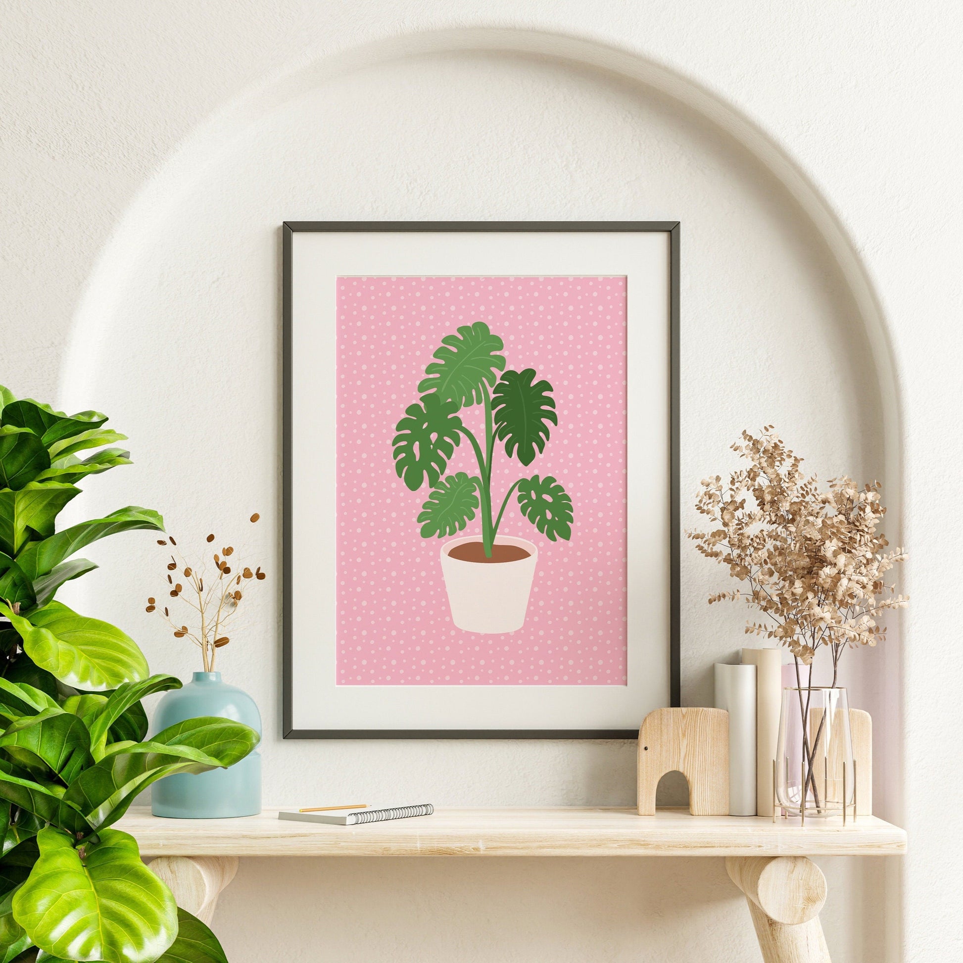 A framed print of a Monstera plant in a pot, with green leaves on a pink background.